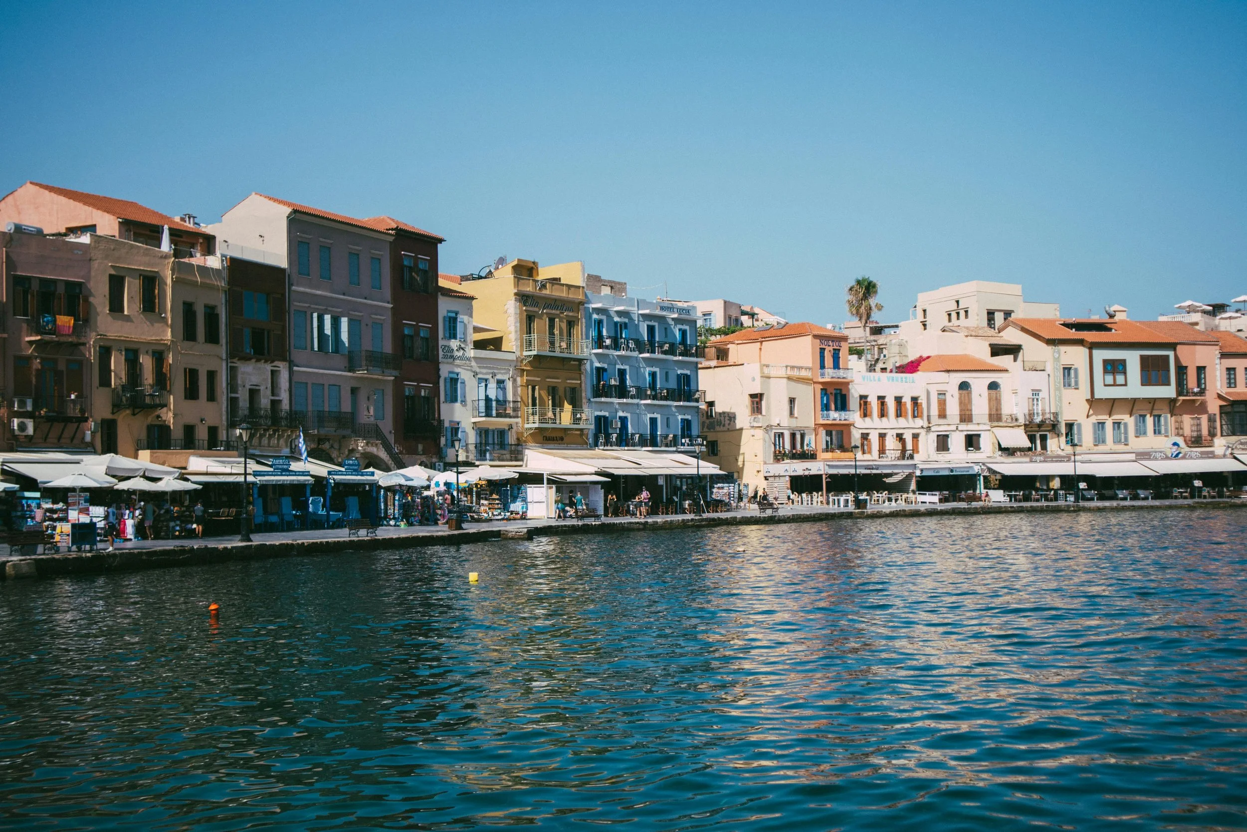 Colorful buildings along a waterfront with shops and cafes, with water in the foreground and a clear blue sky above.