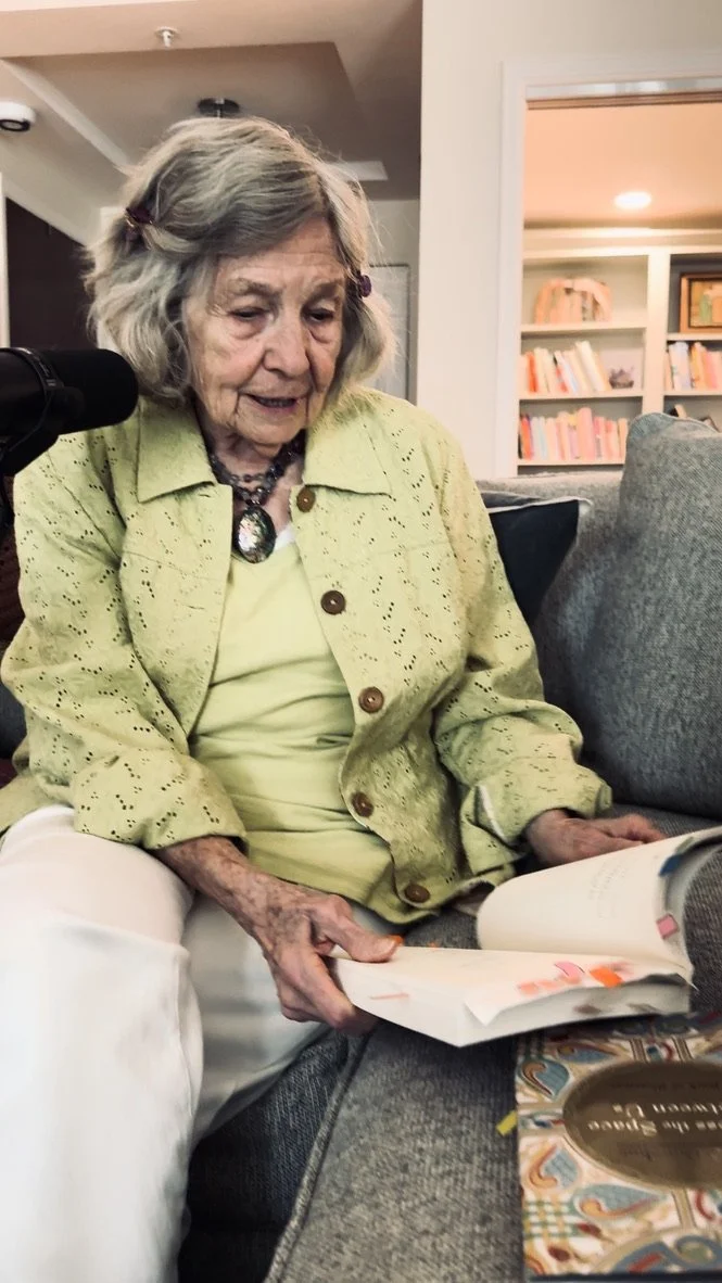 An elderly woman with gray hair and fair skin, wearing a yellow-green embroidered jacket and white pants, is sitting on a couch and reading a book with colorful illustrations.