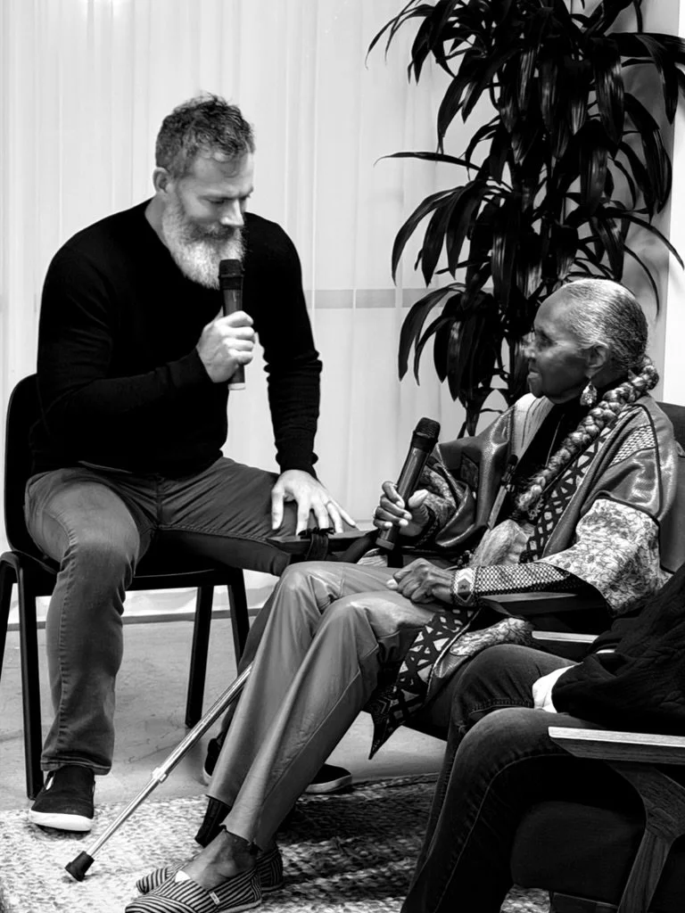 A man with a beard and curly hair sitting on a chair holding a microphone, talking to an elderly woman with braided hair and traditional earrings, who is also holding a microphone and sitting in a chair. There is a large plant behind them.