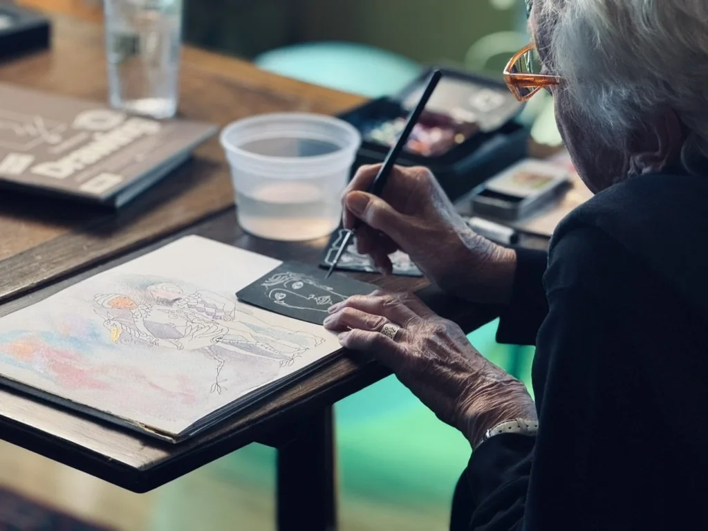 An elderly woman with gray hair and glasses is painting or drawing on paper, seated at a wooden table with art supplies, including a water container, a small black palette, and paper with colorful sketches.