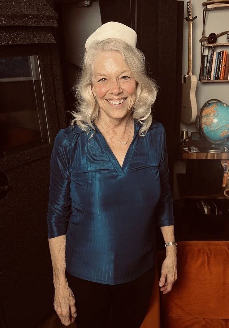An elderly woman with gray, curly hair smiling, wearing a white nurse's cap and a dark blue satin-like blouse, standing in a cozy room with a fireplace, bookshelf, guitar, globe, and an orange chair.
