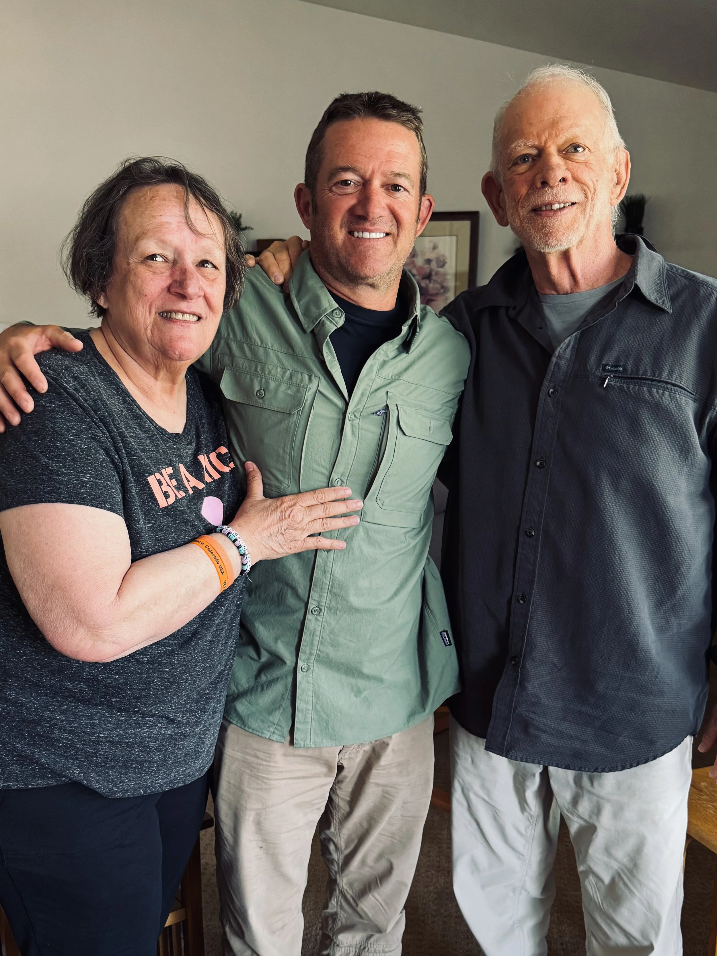 Three smiling people standing close together indoors, arms around each other, posing for a photo.