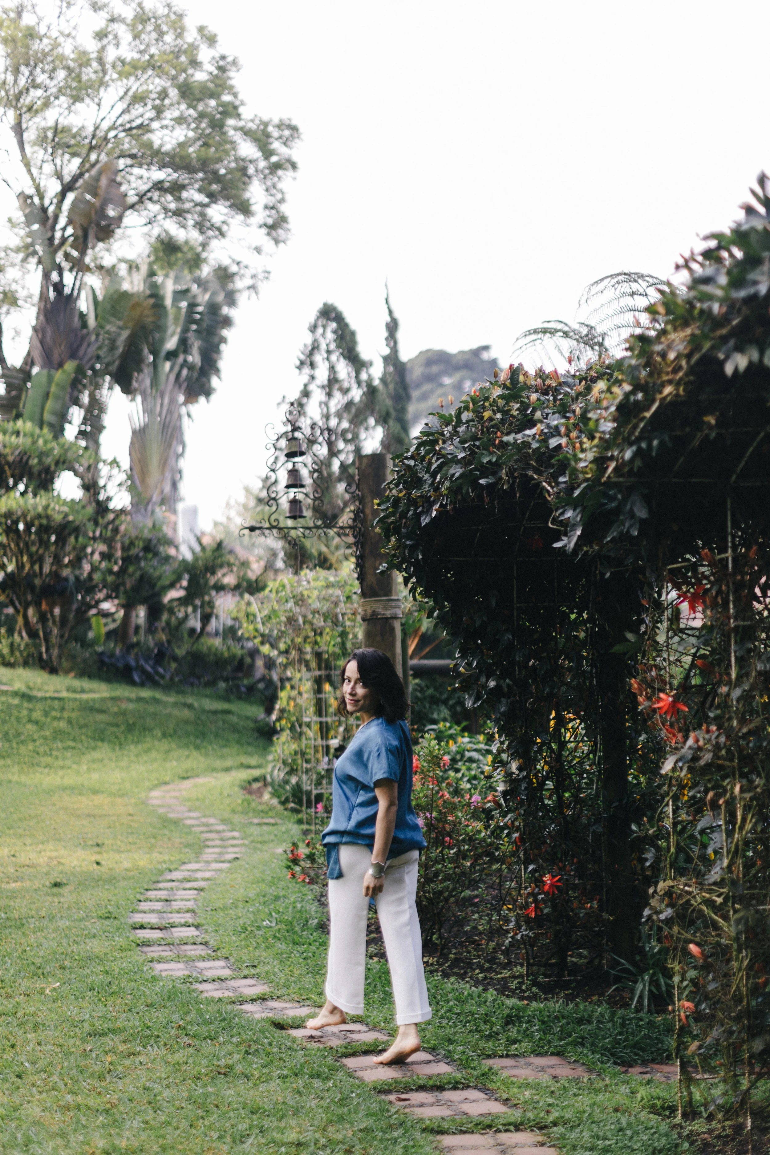 Woman standing on a garden path surrounded by greenery and flowers.