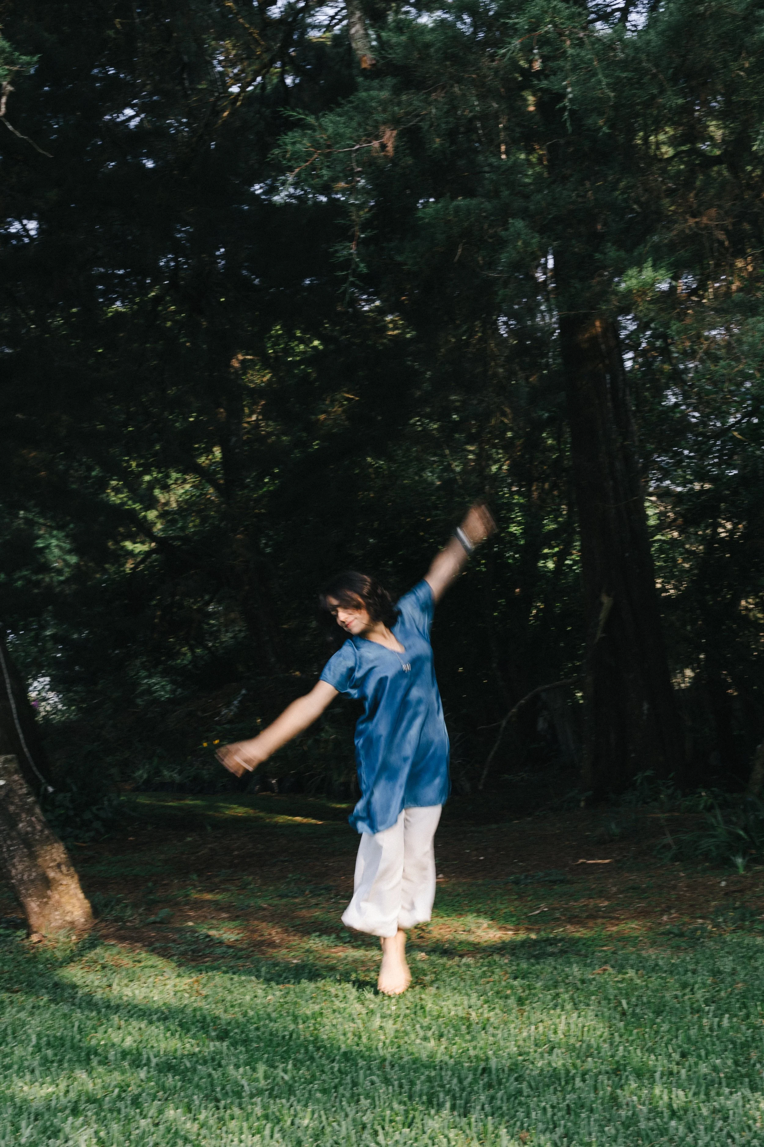 Person in a blue shirt and white pants dancing on grass in a wooded area.