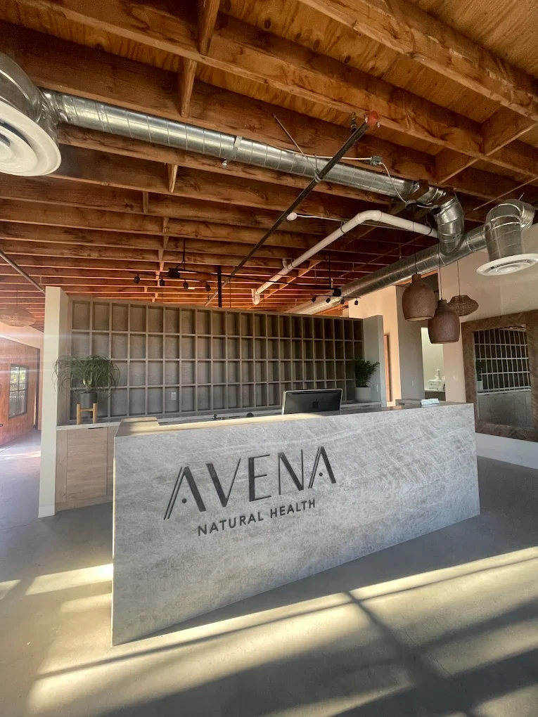Reception desk with the sign 'AVENA NATURAL HEALTH' in a modern interior with wooden ceiling and metal pipes.