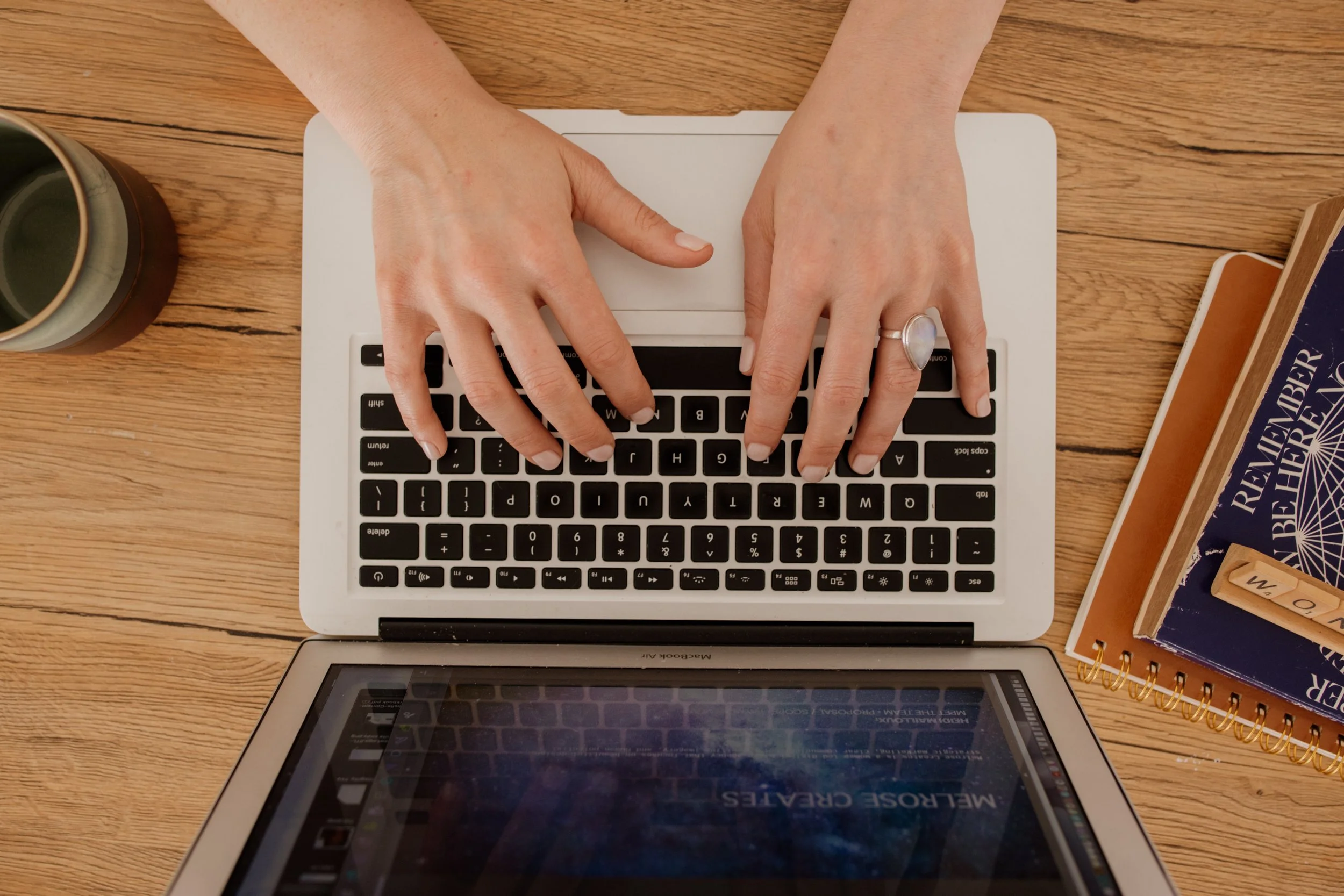 Hands typing on a laptop keyboard on a wooden desk, with a cup and notebooks nearby.