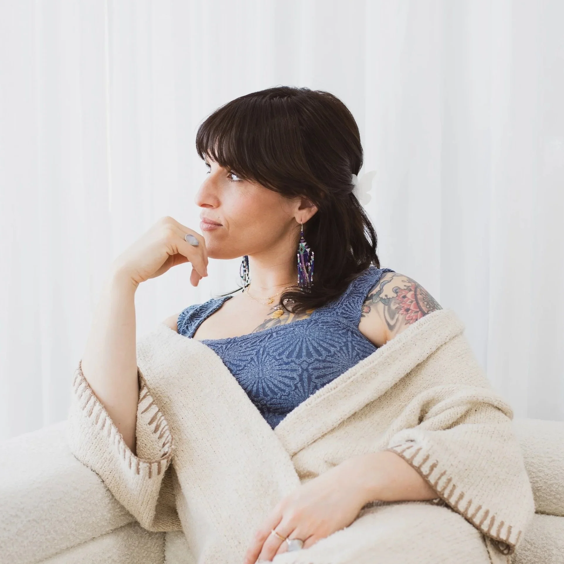 Side profile of a woman with dark hair, wearing a blue top and colorful earrings, sitting on a cream-colored sofa with a cream cardigan draped over her shoulders.