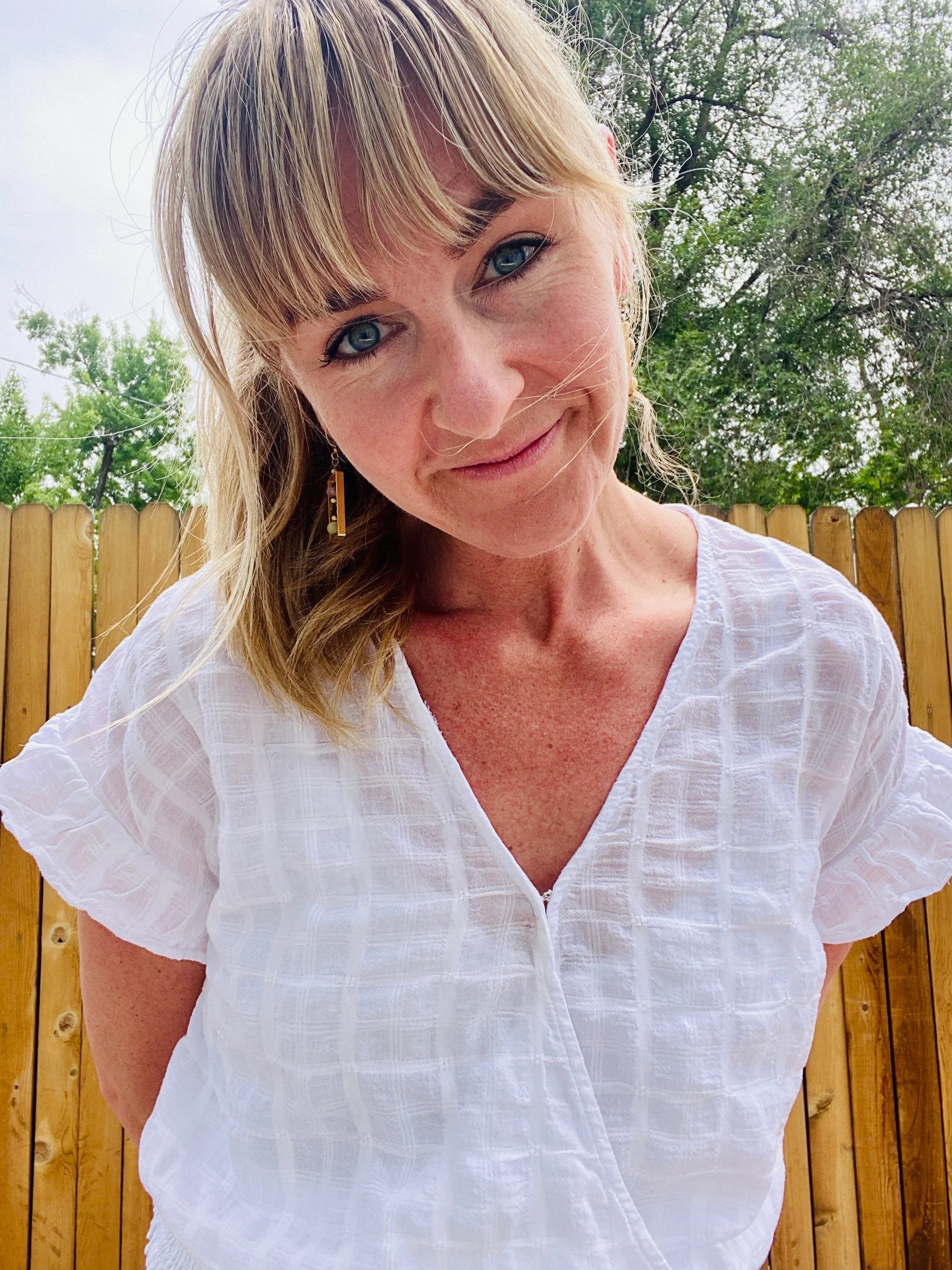 A woman with blonde hair, blue eyes, and light skin, smiling outdoors in front of a wooden fence and green trees, wearing a white blouse.