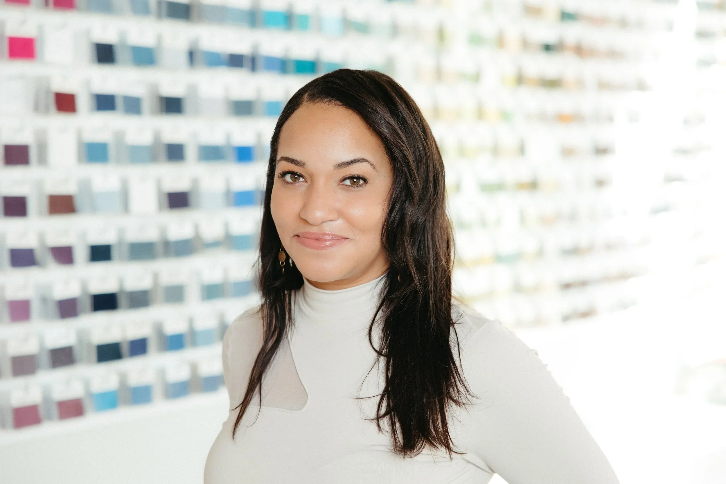 A woman with long dark hair wearing a white top, standing indoors in front of a colorful, patterned background.
