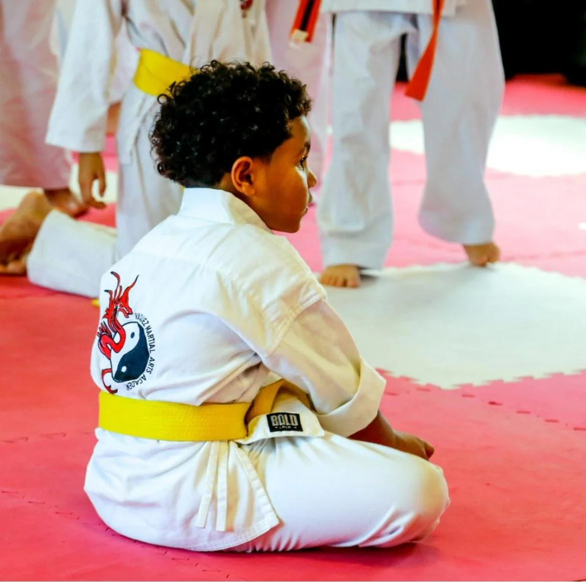 A young boy with curly hair sitting on a red mat in a martial arts dojo, wearing a white uniform with a yellow belt, attentively watching a martial arts class in session around him.