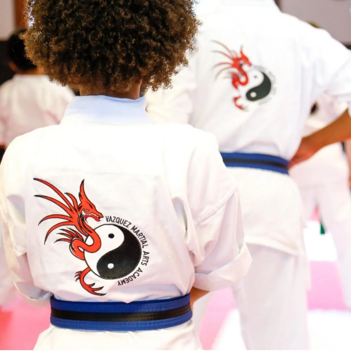 Martial arts students wearing white uniforms with a dragon and yin-yang logo on the back, standing in a training area.