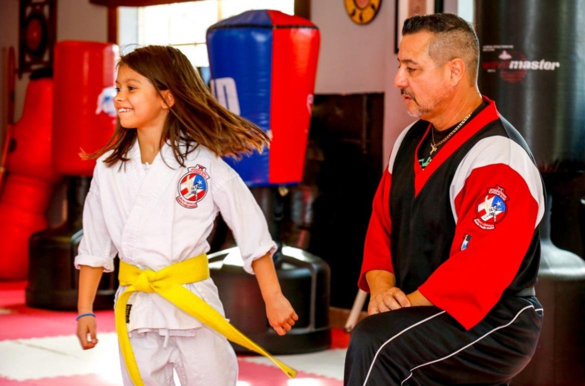 A young girl in a karate gi with a yellow belt smiling and moving, while a male instructor sits nearby observing, in a martial arts training center with punching bags in the background.