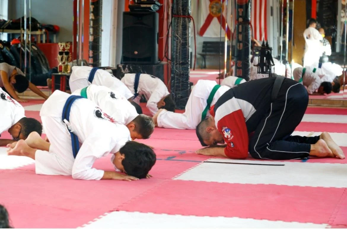 Martial artists bowing during a class or practice session in a dojo or gym with mirrors, flags, and training equipment.