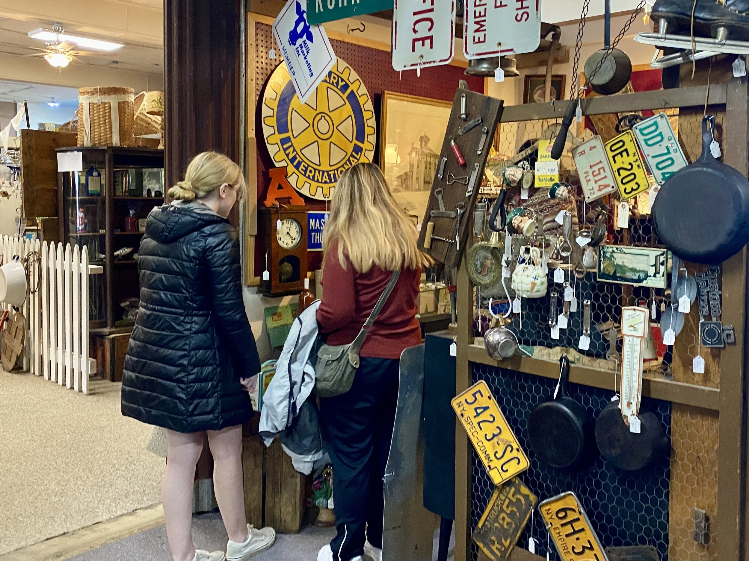 Two people browsing vintage items and antique signs in a shop filled with old tools, cookware, and collectibles.