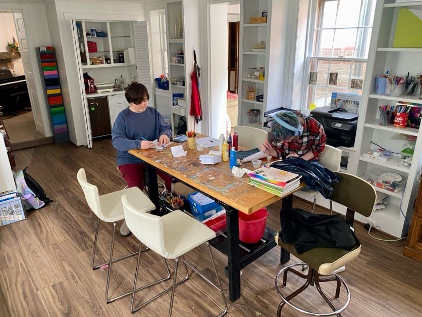 Two children focused on creative activities at a cluttered wooden table in an art studio, demonstrating authentic self-directed learning through exploration and personal interest.
