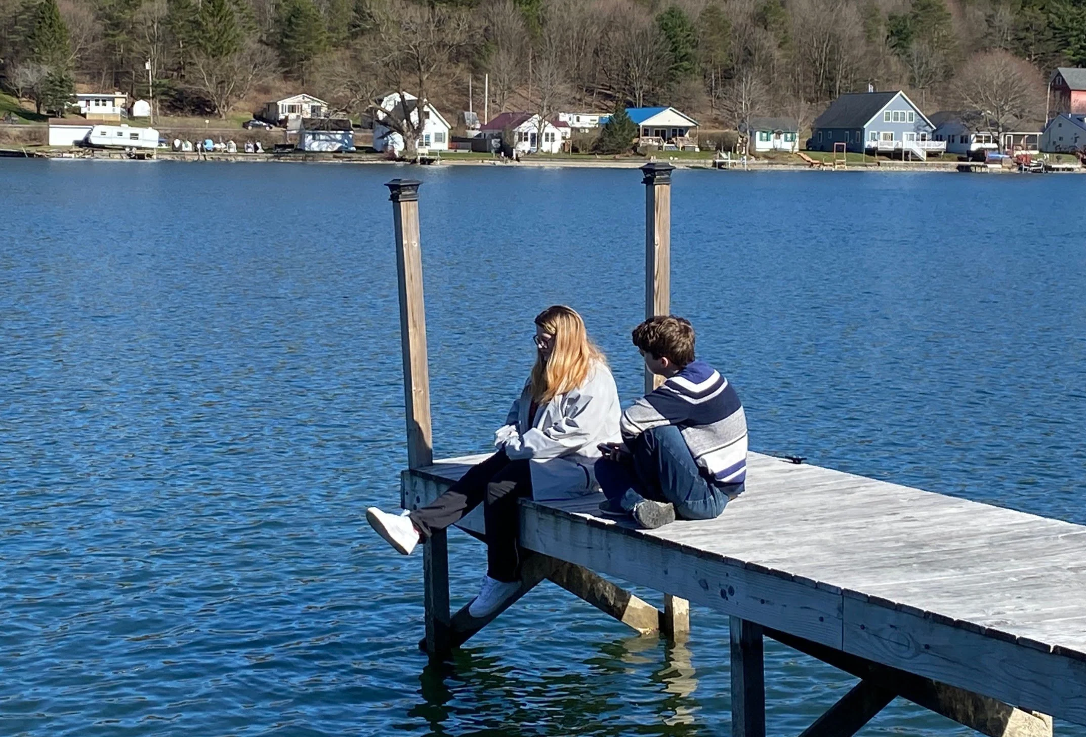 Two teens sitting on a dock near a lake.