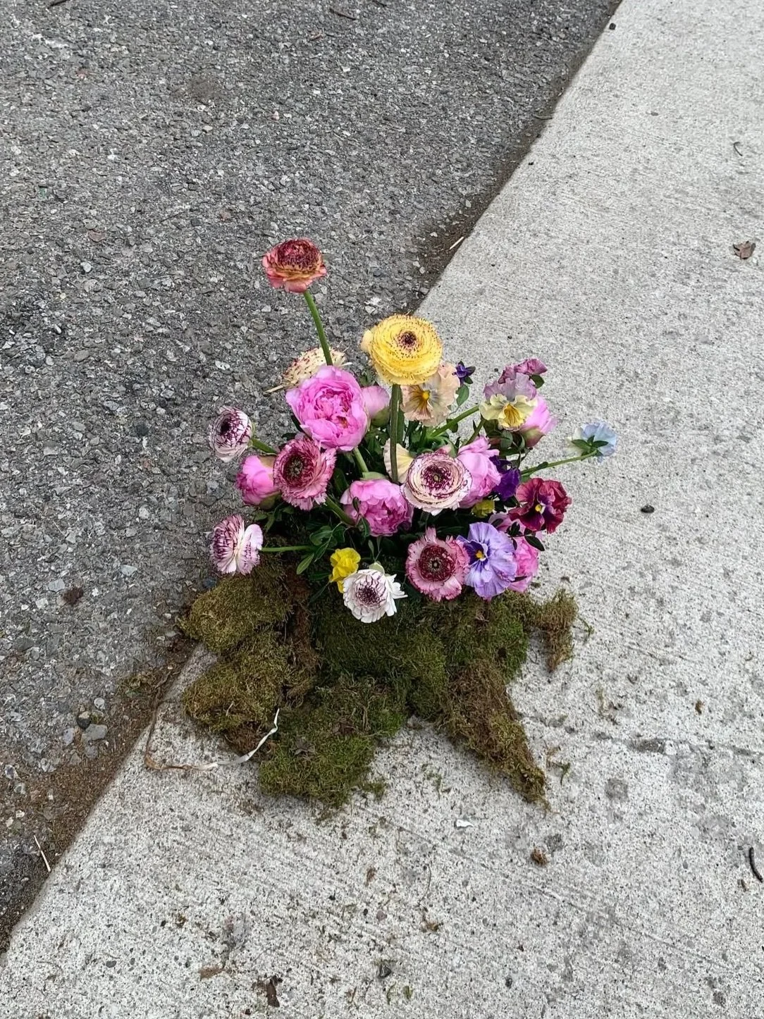 A small bouquet of colorful flowers, including pink, purple, and yellow blossoms, arranged on a patch of moss at the corner of a sidewalk and asphalt street.