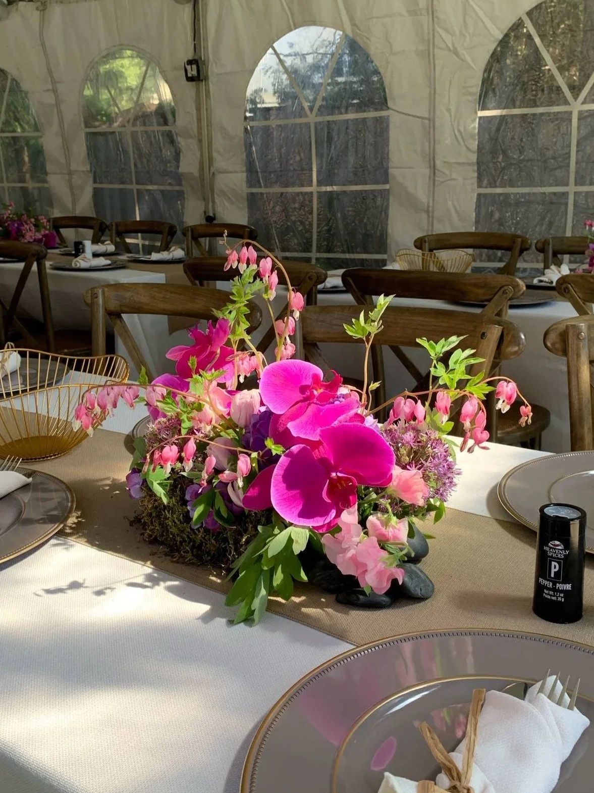 A table decorated with a pink floral centerpiece in a tented outdoor dining area with wooden chairs and a white tablecloth.