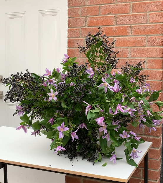 Large floral arrangement with purple and pink flowers, green leaves, and dark berries on a white table against a brick wall and white door.