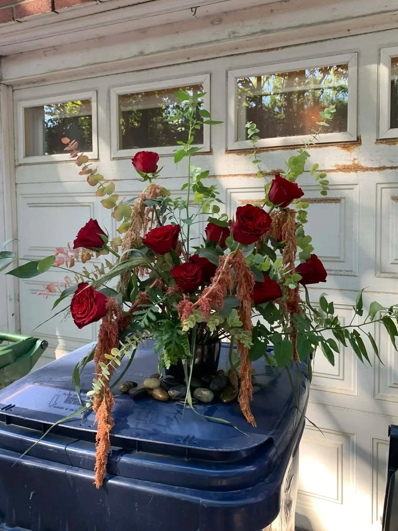 A floral arrangement with red roses, various foraged greens, and hanging amaranthus in a black vase.