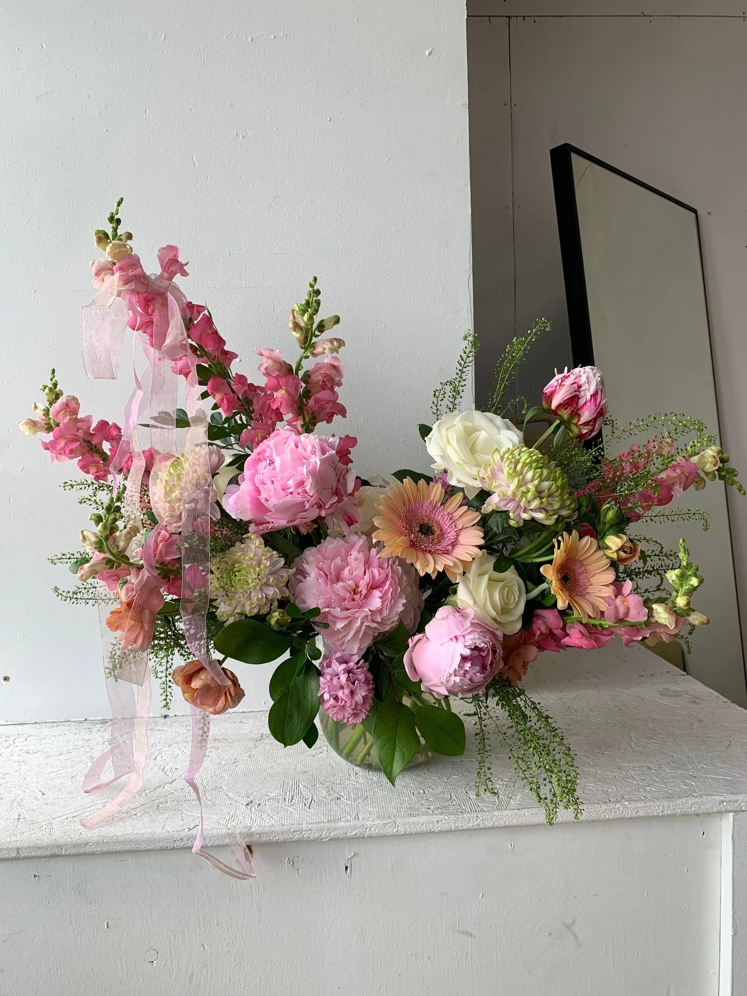 A floral arrangement with pink, white, and peach flowers, including roses, peonies, and gerbera daisies, decorated with a pink ribbon, placed on a white surface against a plain background.