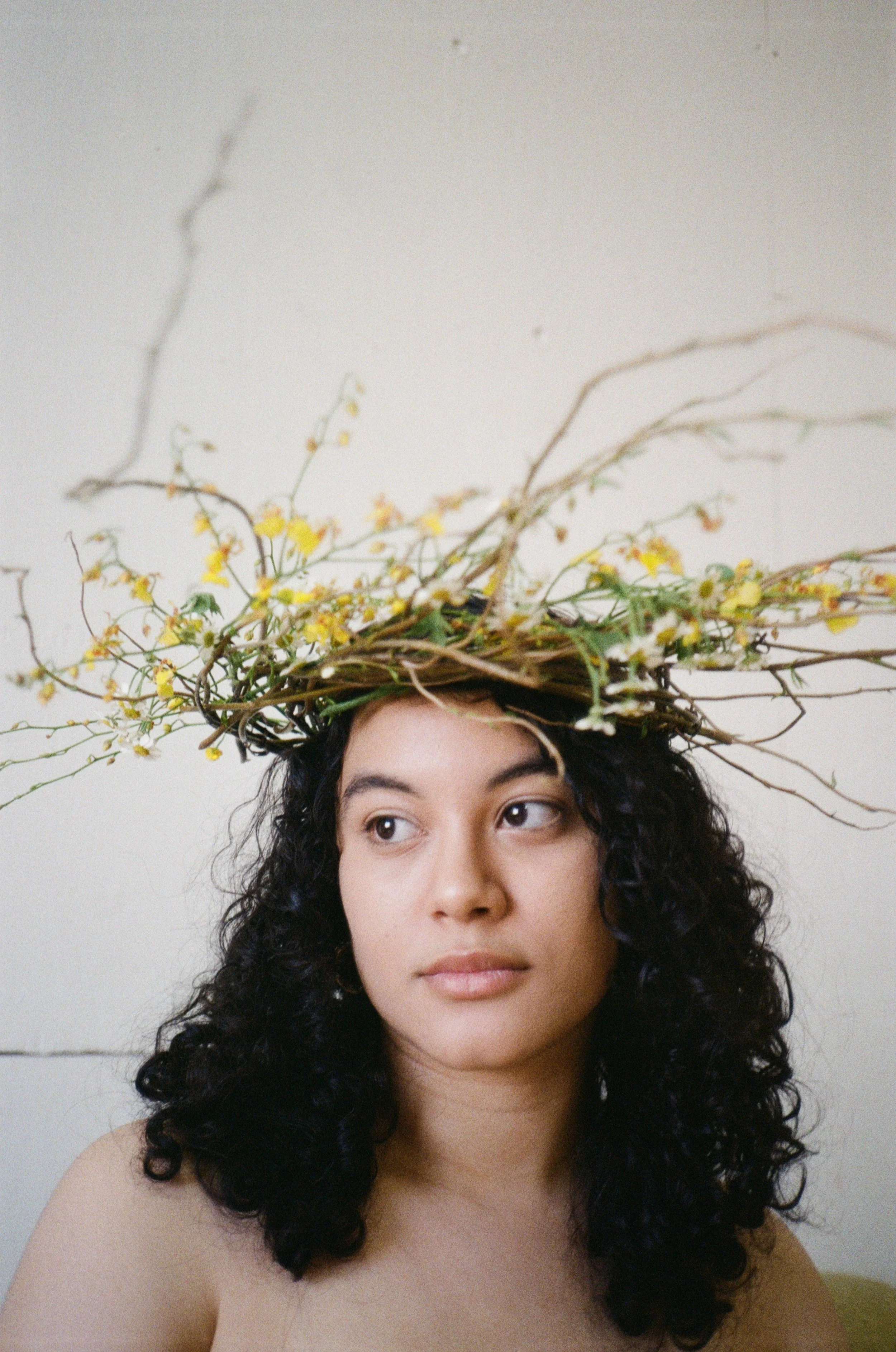 A woman with dark curly hair wearing a crown made of twigs and yellow flowers.