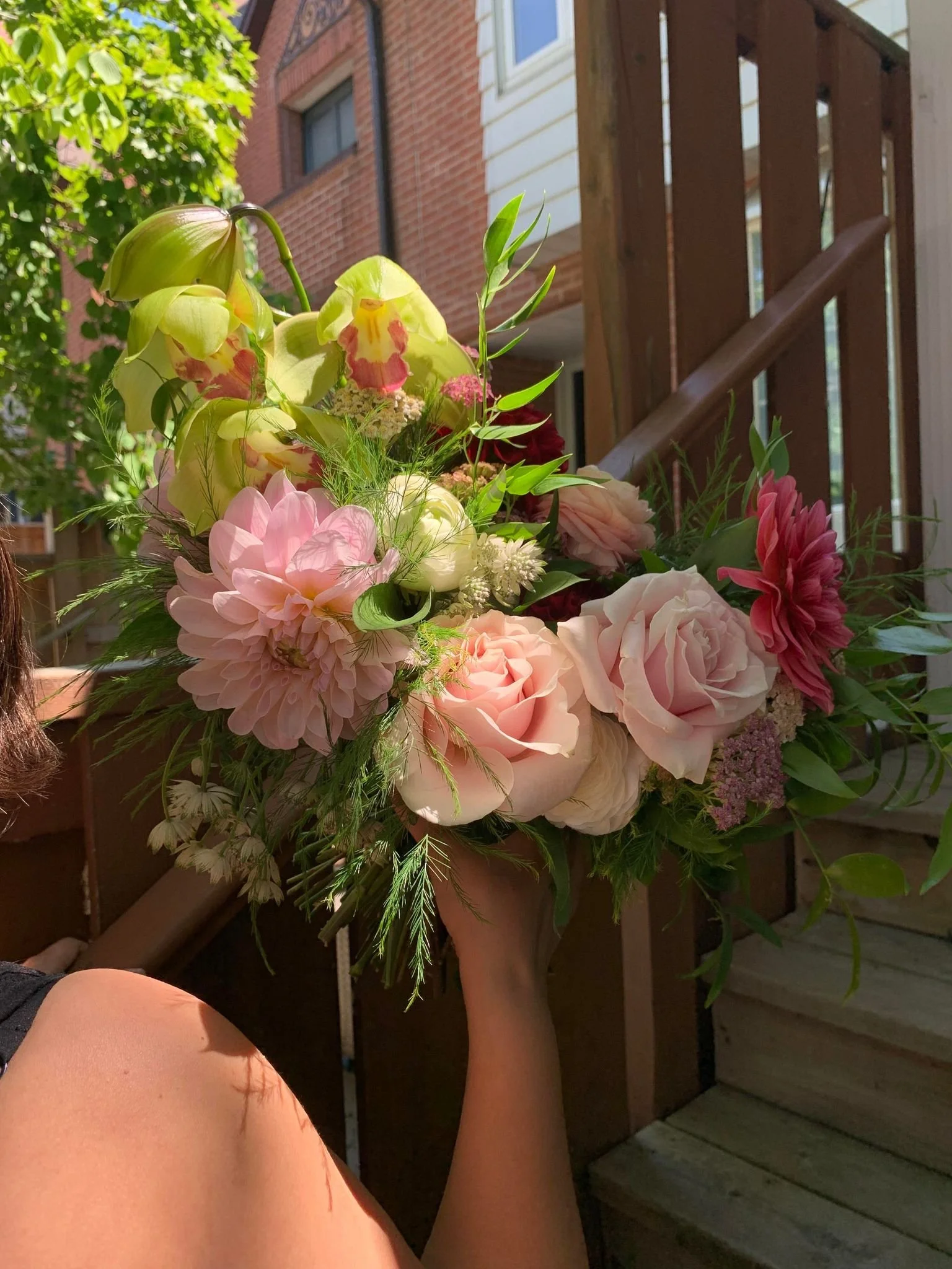 A person holding a bouquet of mixed flowers including pink roses, dahlias, and other blooms, outdoors.