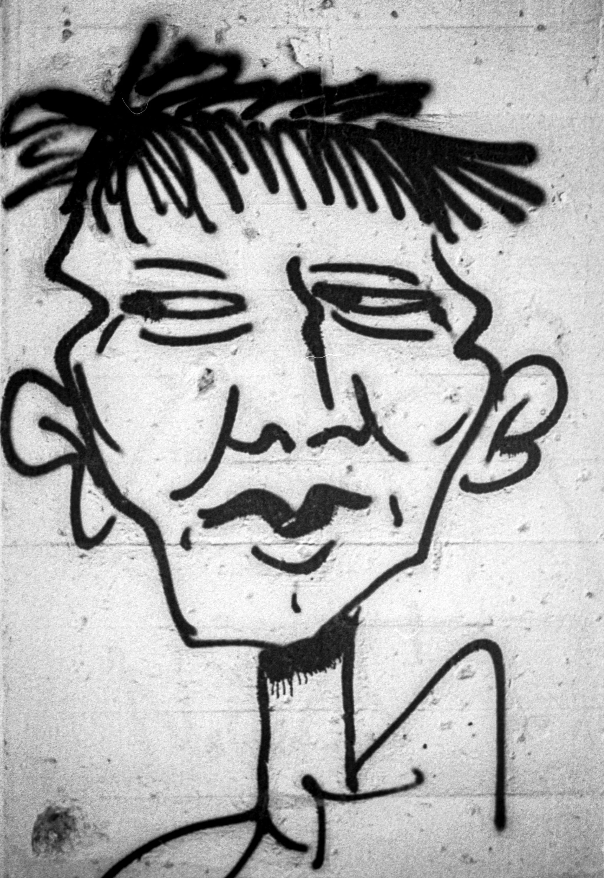 Black and white graffiti art of a person's face with exaggerated features, including prominent ears, messy hair, and a mustache, drawn with bold lines on a textured wall.