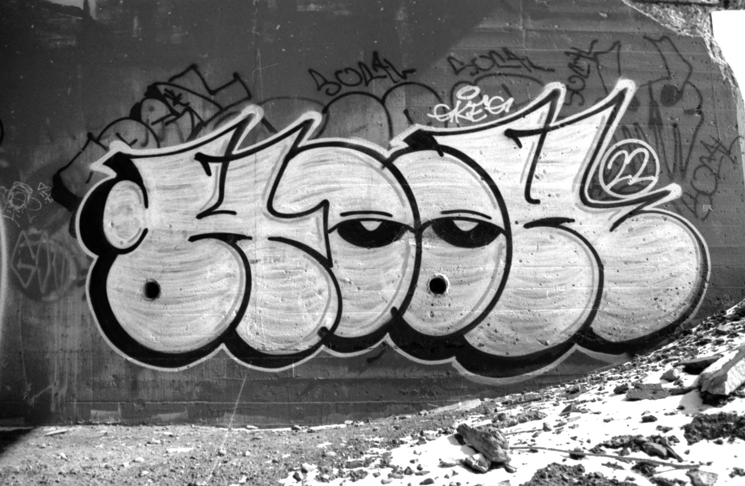 Black and white graffiti on a concrete wall featuring the word 'SCOOB' with the letter 'O' depicted as a pair of eyes with droopy eyelids. The wall is surrounded by smaller tags and graffiti art, and there are rocks and snow on the ground in front of