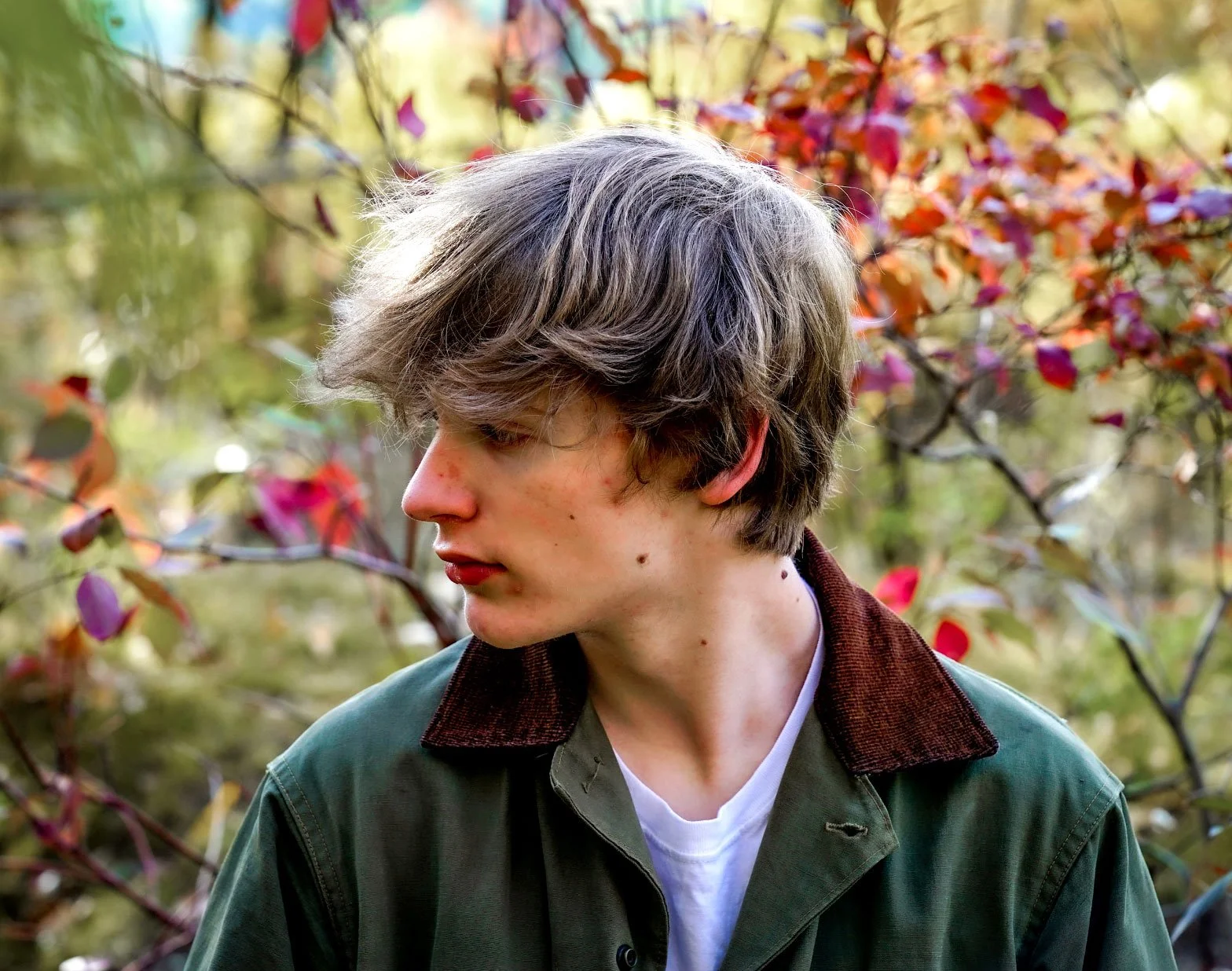 A young man with shaggy gray hair and fair skin, wearing a green jacket with a brown collar, standing outdoors among autumn-colored bushes.