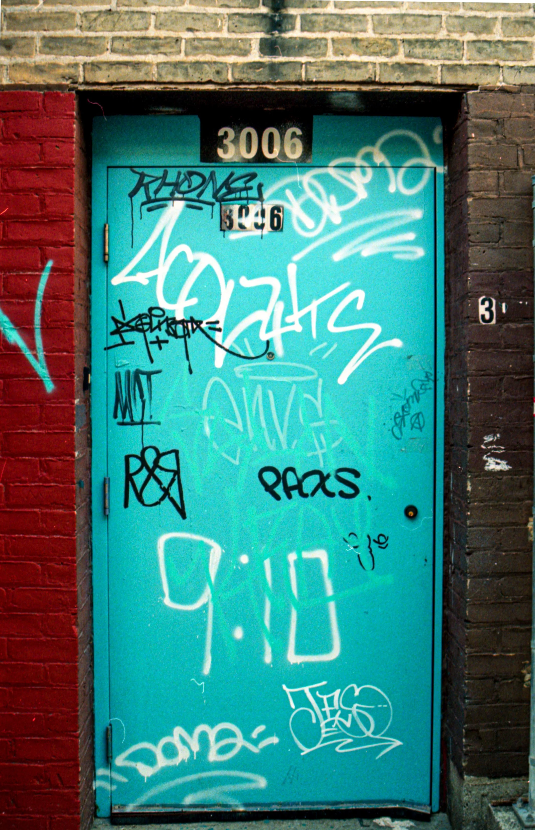 A bright blue door covered in various black and white graffiti tags, set within brick walls painted red on the left and black on the right.