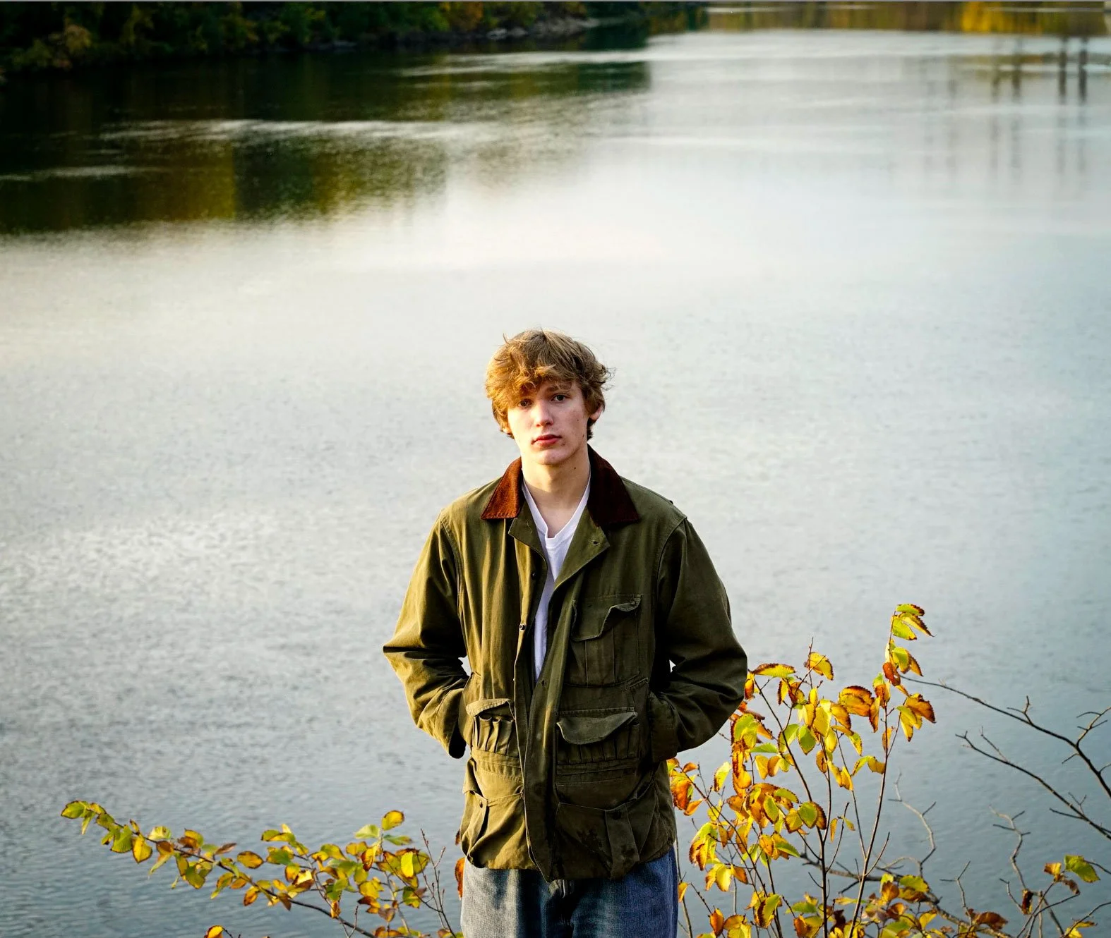 A young man with curly brown hair wearing a green jacket and blue jeans standing by a body of water with trees and branches with yellow and green leaves in the background.