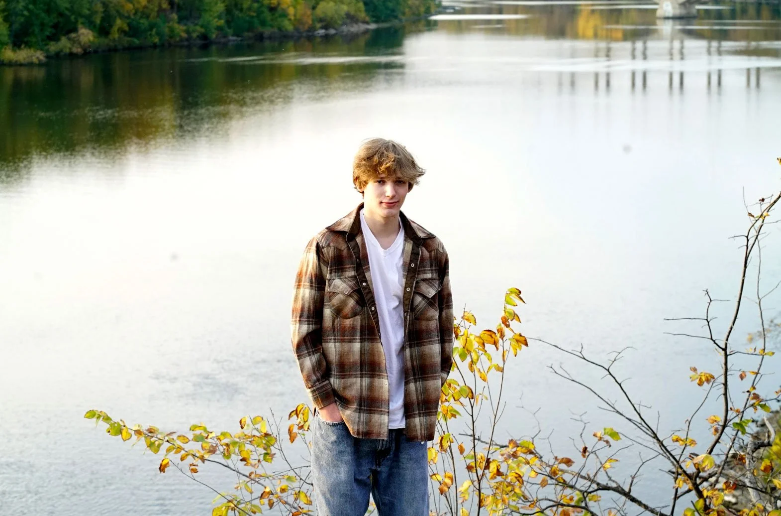 A teenage boy standing outdoors near a body of water during autumn, wearing a plaid jacket over a white shirt and jeans.