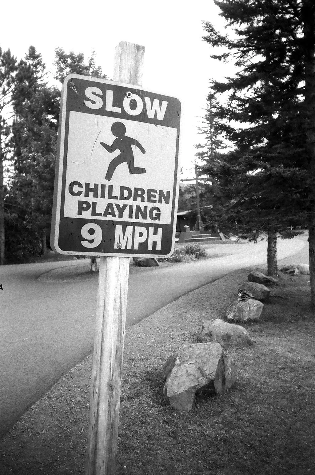 A black and white photograph of a road sign that reads "Slow Children Playing 9 MPH" with a silhouette of a child. The sign is mounted on a wooden post next to a curved road, with trees and rocks along the roadside.