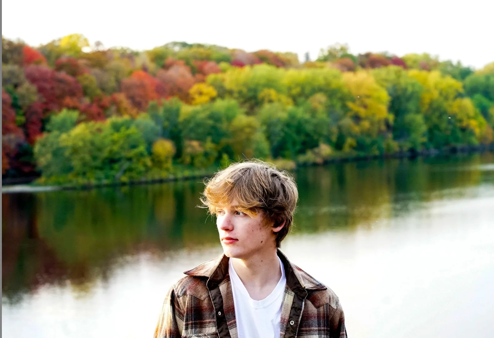 A young man with light brown, slightly wavy hair stands outdoors near a body of water, with colorful autumn trees in the background.