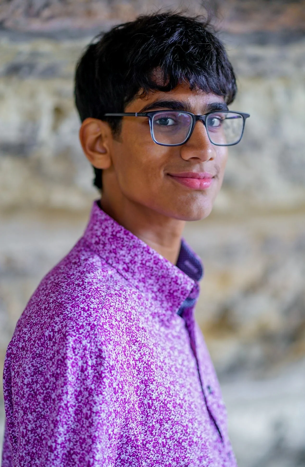 Portrait of a young man with glasses wearing a purple patterned shirt against a stone wall background.
