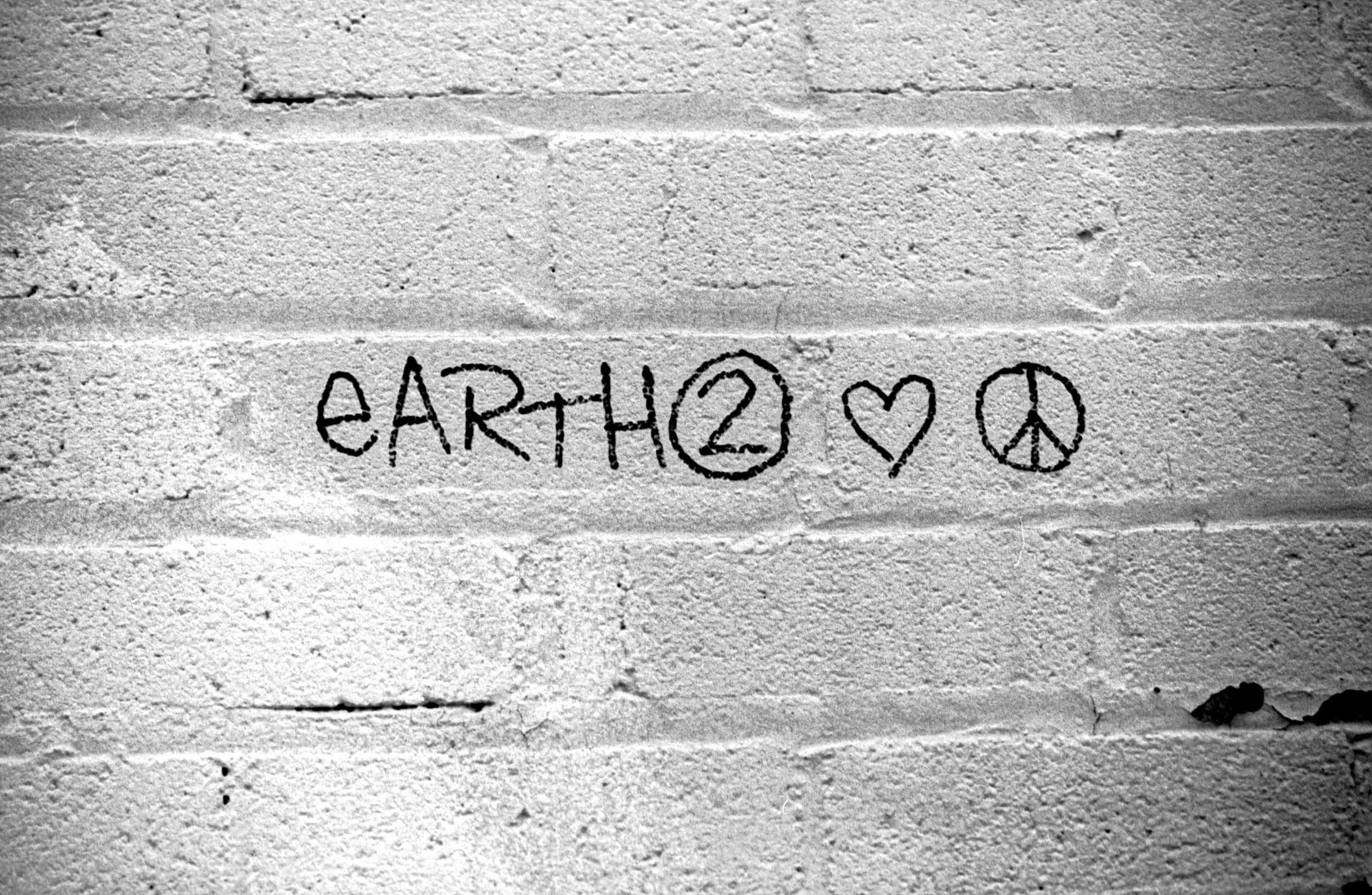 Graffiti on a white brick wall reading 'eARTH2' with a circle around the '2', a heart, and a peace symbol.