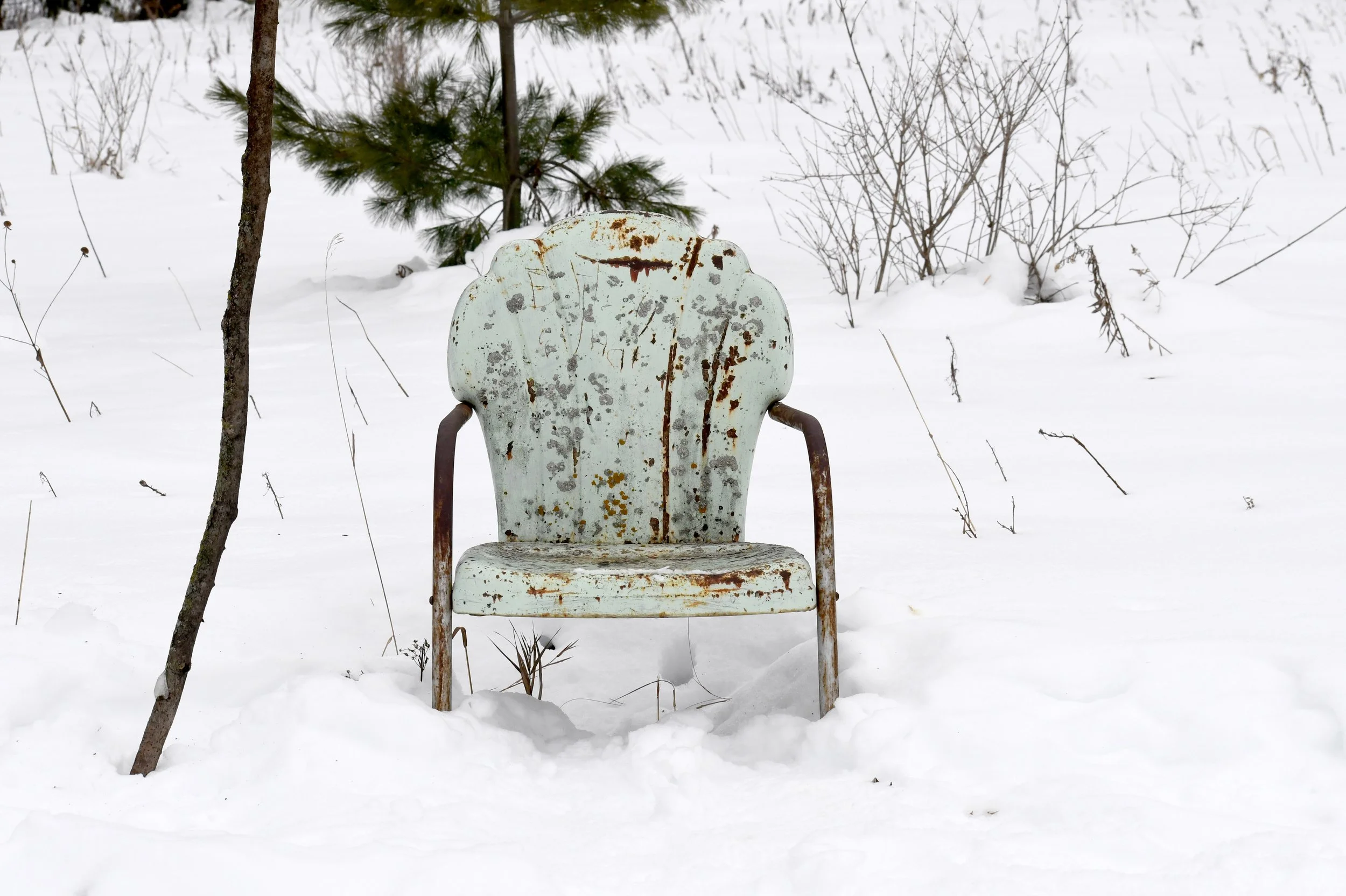 An abandoned, rusty metal chair with a faded light greenish color is placed in a snowy field surrounded by leafless bushes and trees.