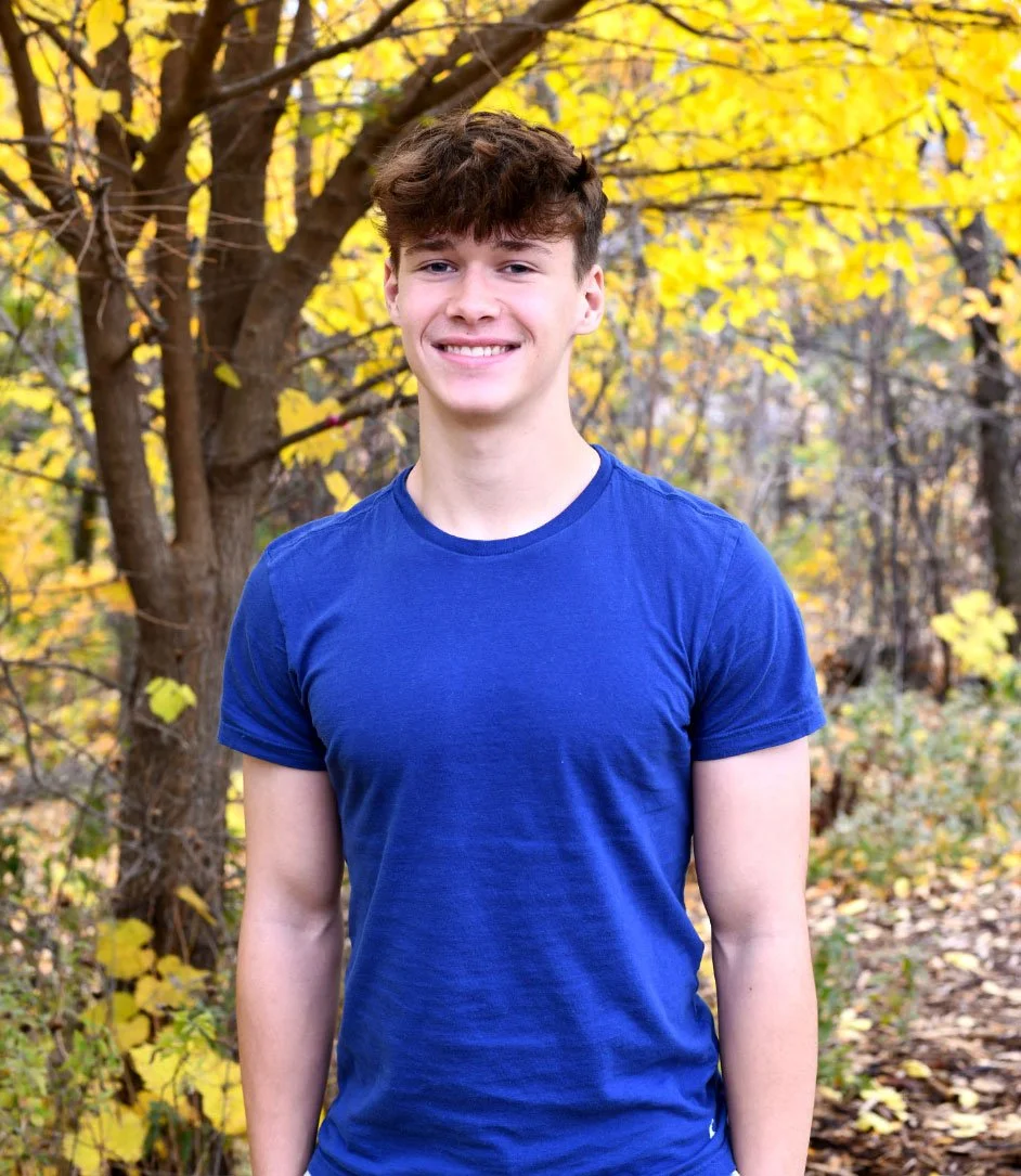 A young man with brown hair wearing a blue T-shirt standing outdoors in a forest with yellow autumn leaves.