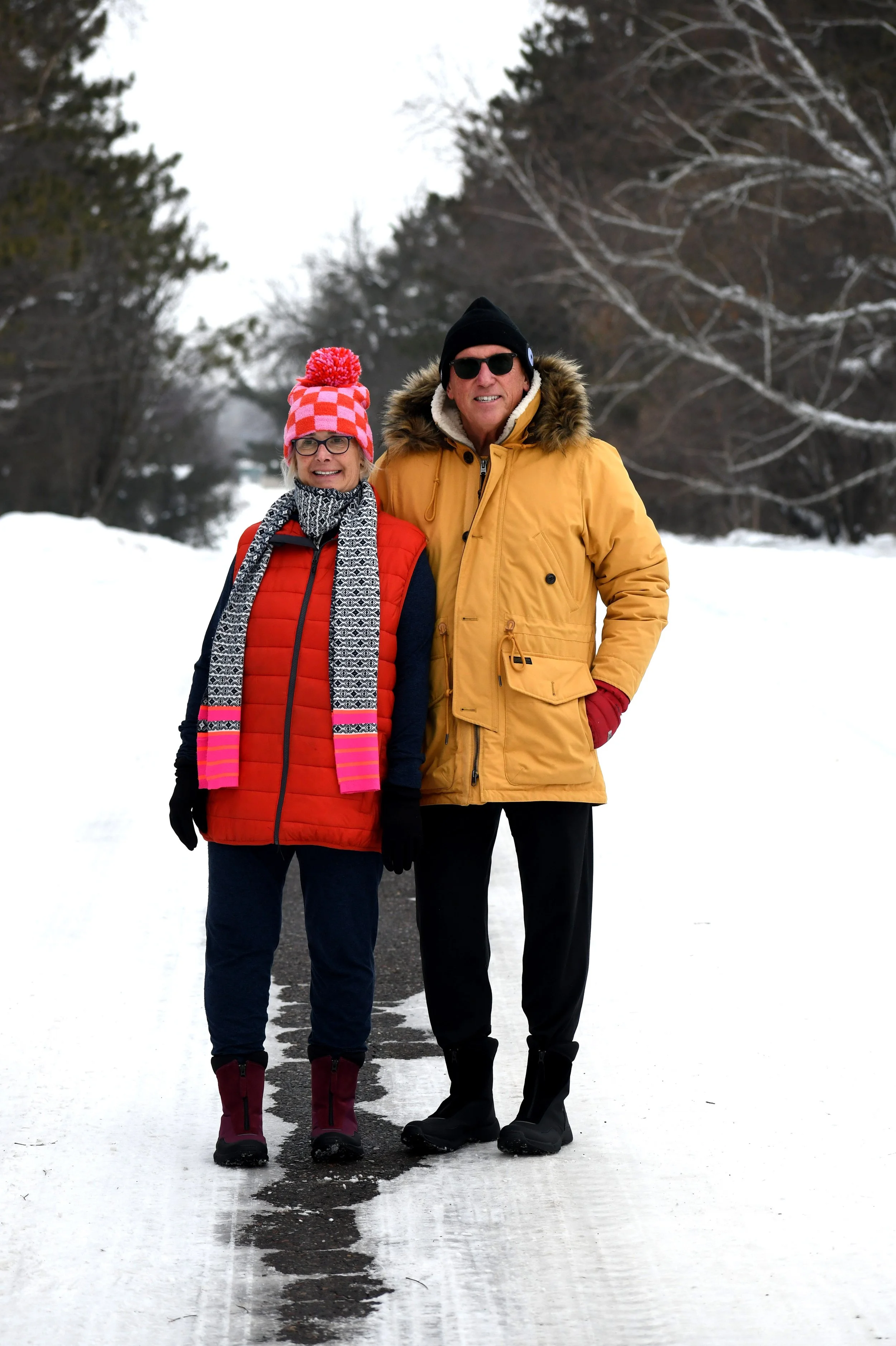 An elderly man and woman stand together outdoors on a snowy path, dressed in winter clothing, smiling at the camera.