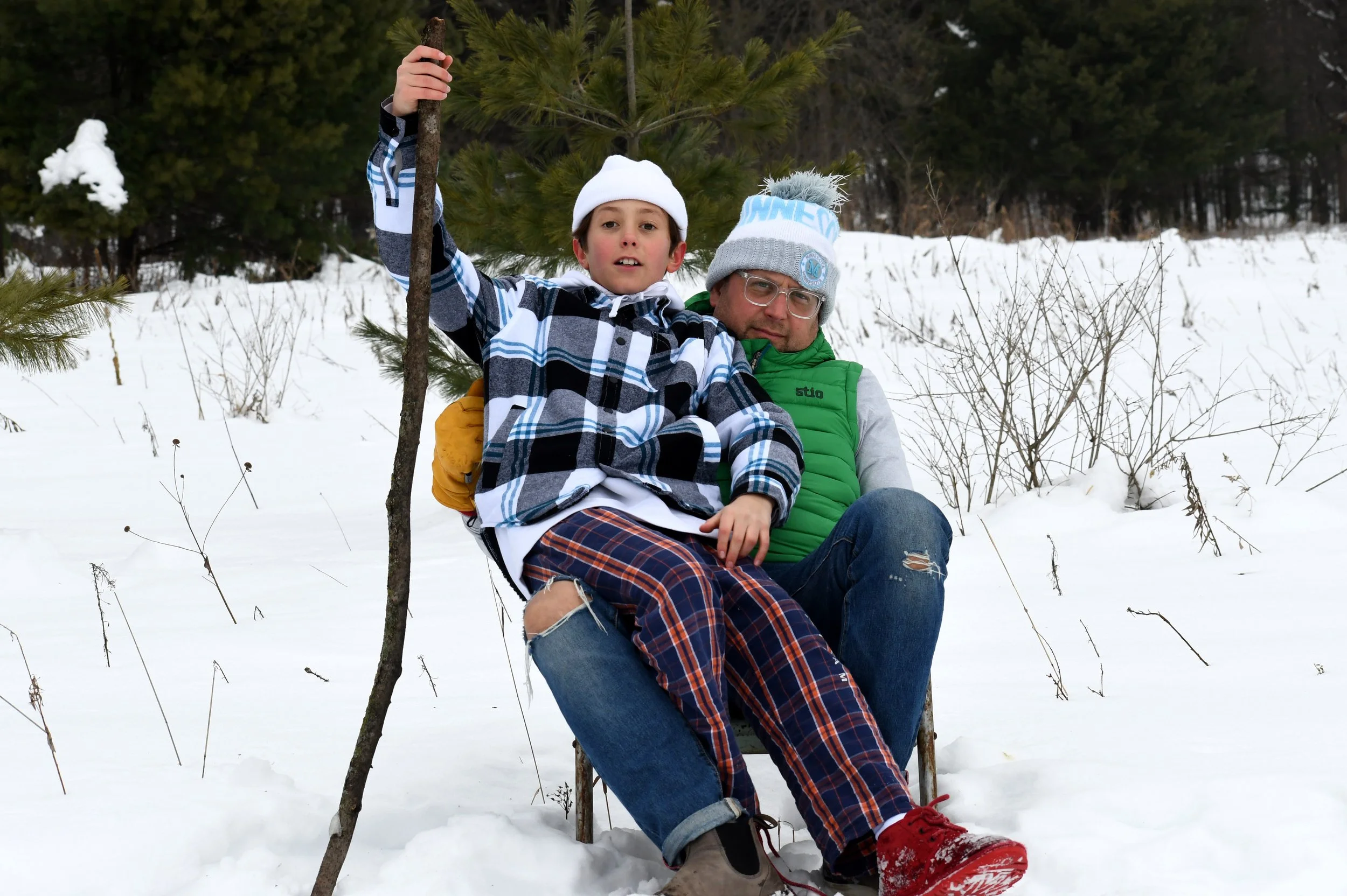 A man and a boy sitting on a sled in a snowy forest, wearing winter clothes and hats. The boy holds a stick.