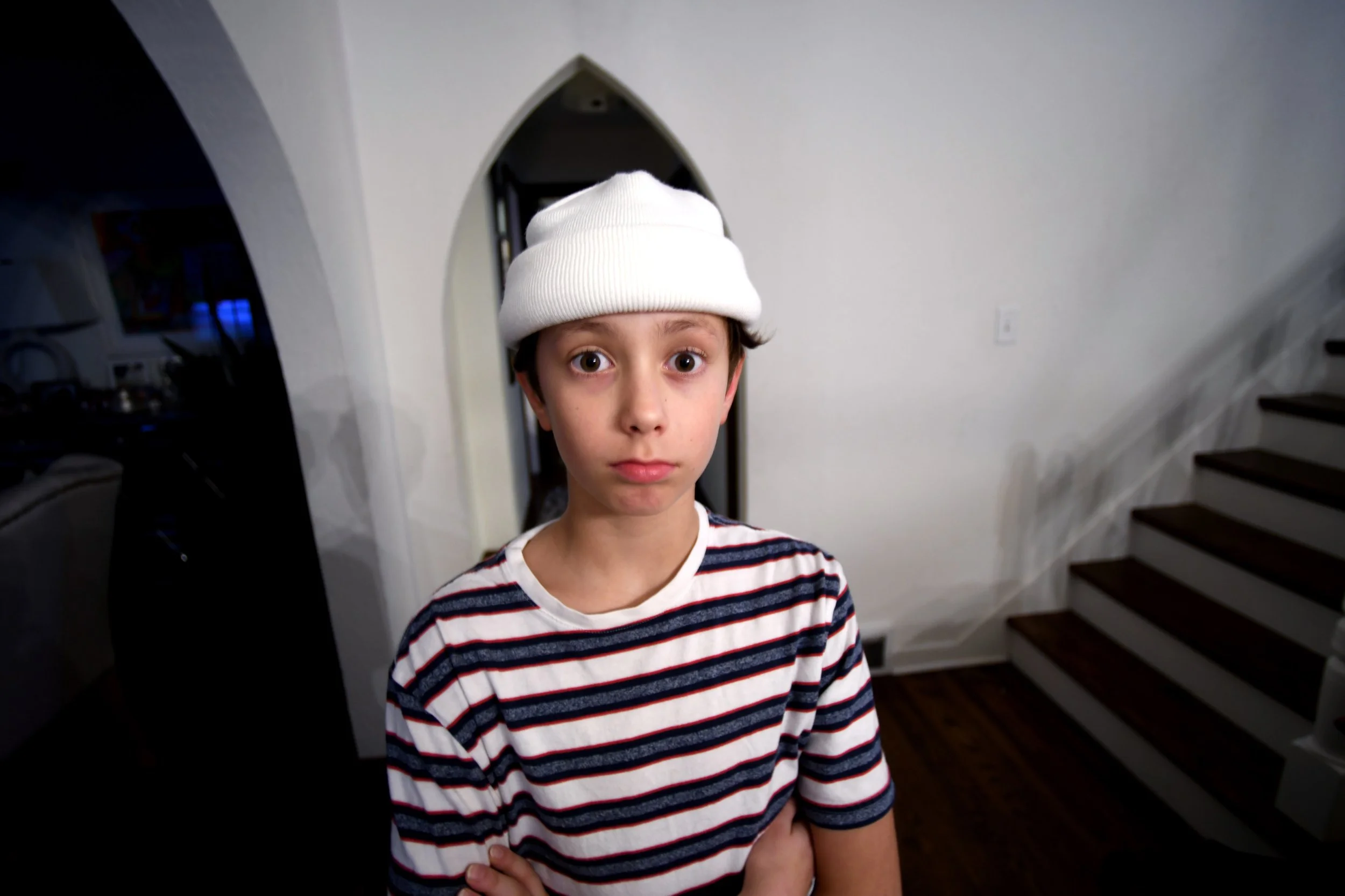 A young boy wearing a white beanie and a striped t-shirt standing with crossed arms inside a house, looking directly at the camera with a neutral expression.
