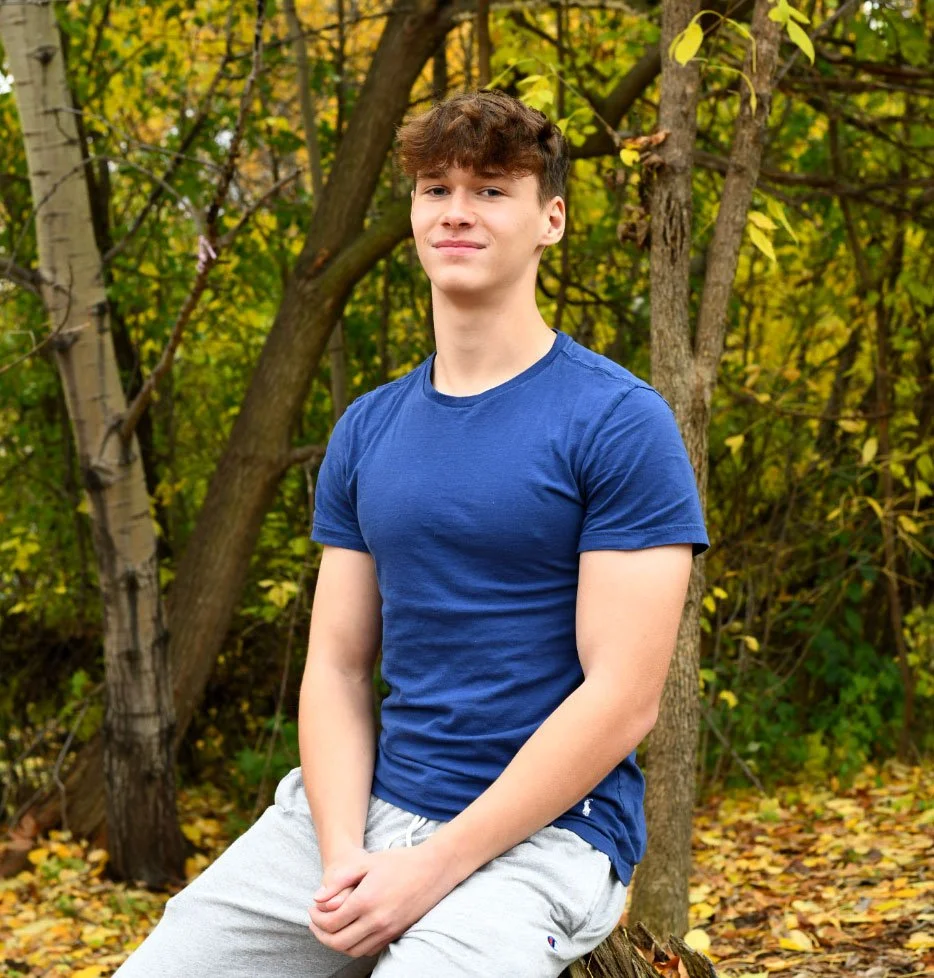A young man sitting outdoors in a forested area with yellow and green autumn leaves, wearing a blue t-shirt and light gray shorts, looking at the camera.