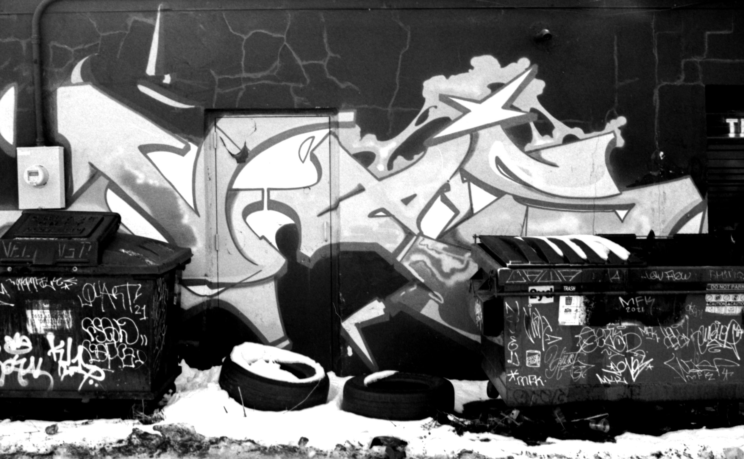 Street graffiti wall with two graffiti-covered dumpsters and snow on the ground.