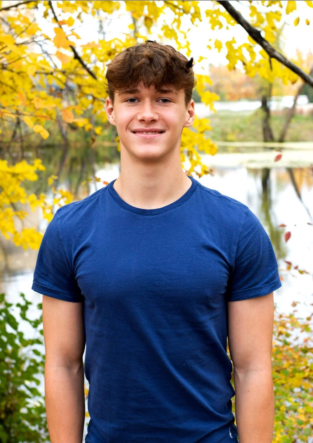 A young man wearing a blue t-shirt standing outdoors by a lake with yellow autumn leaves and trees in the background.
