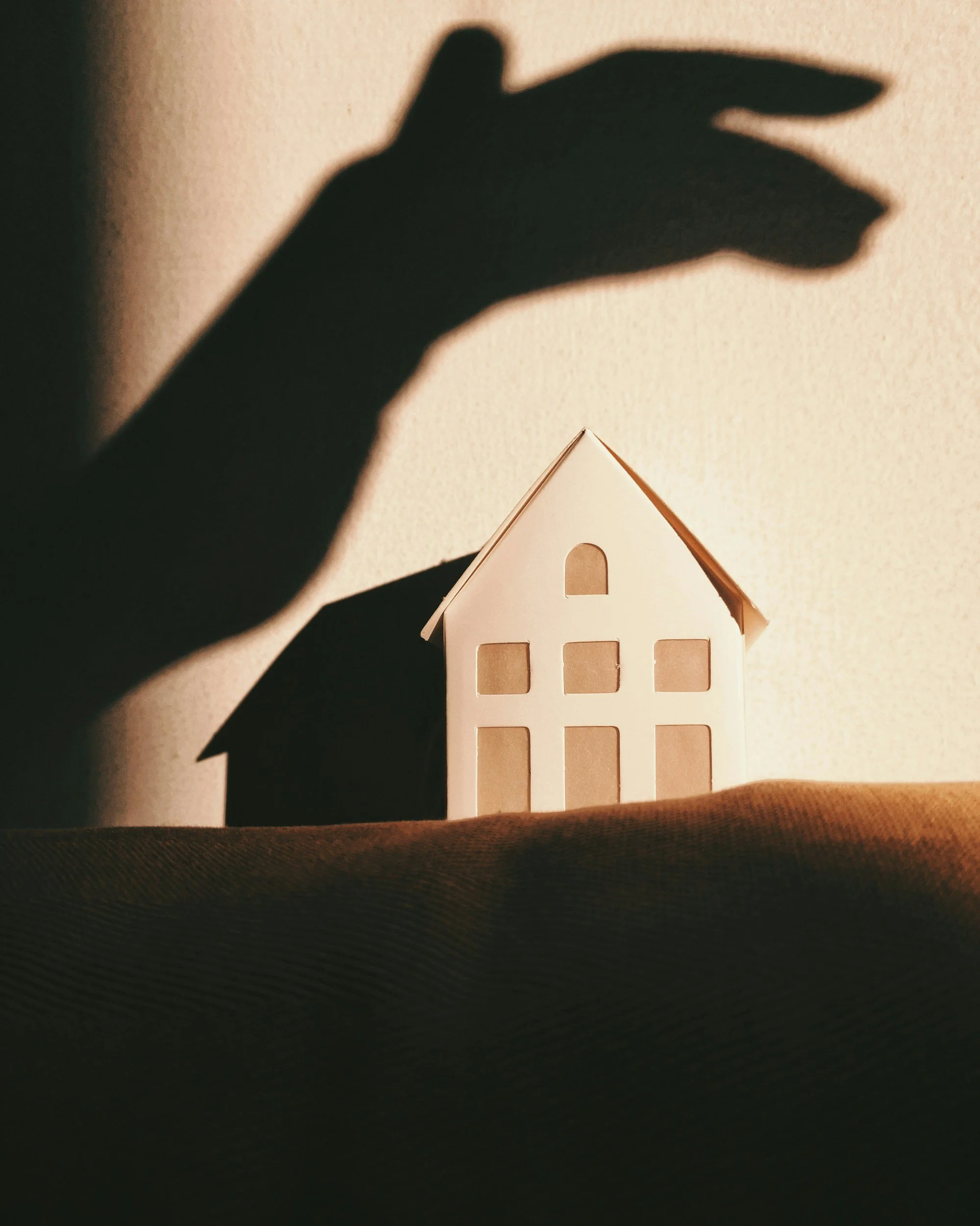 Shadow of a hand cast on a paper house with cutouts, lit from the side, creating a silhouette against a neutral background.