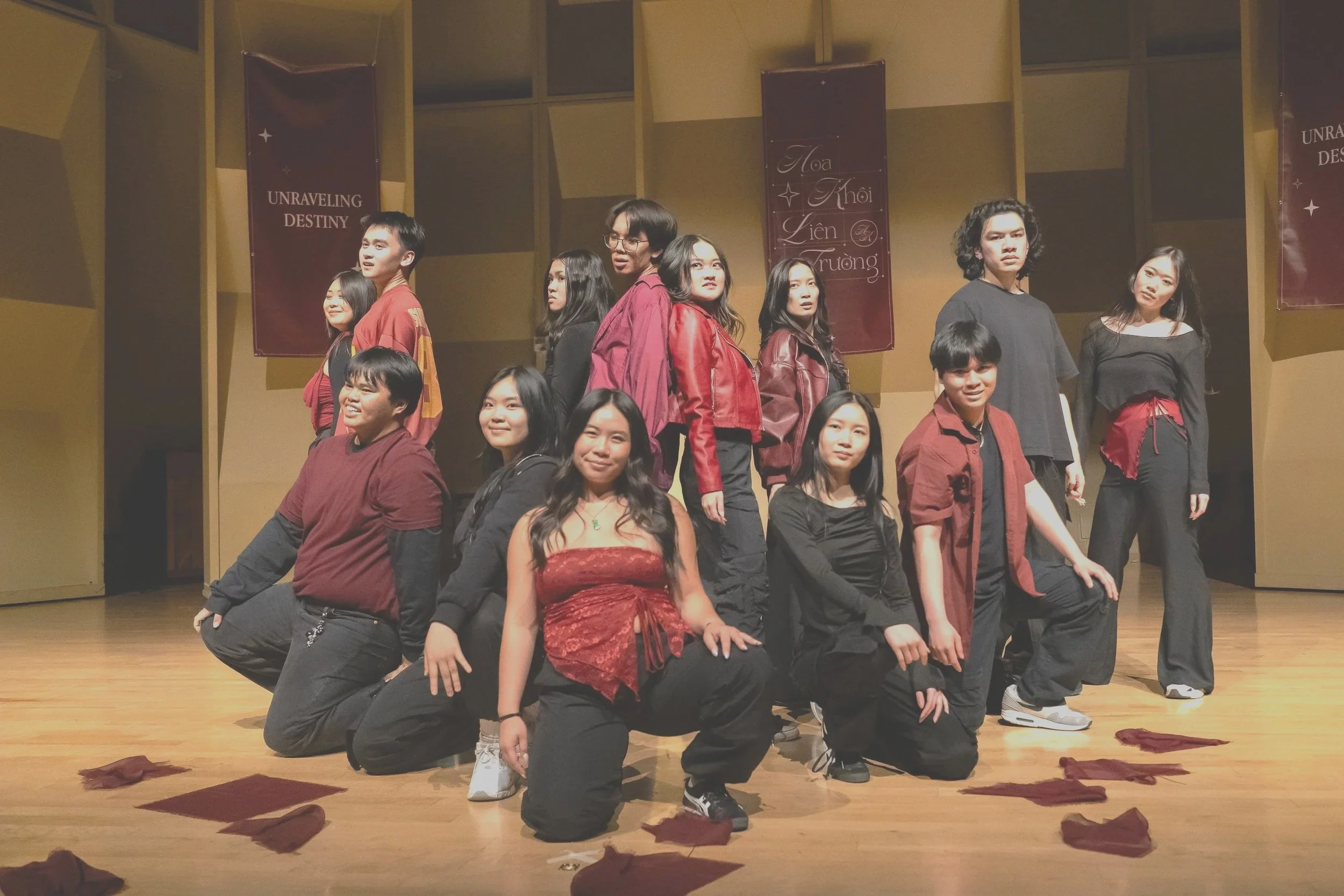 Group of young people posing on stage, some kneeling and others standing, during a performance or event. The background has banners with text including 'Unraveling Destiny' and Vietnamese words. The stage has scattered fabric pieces and a wooden floor.