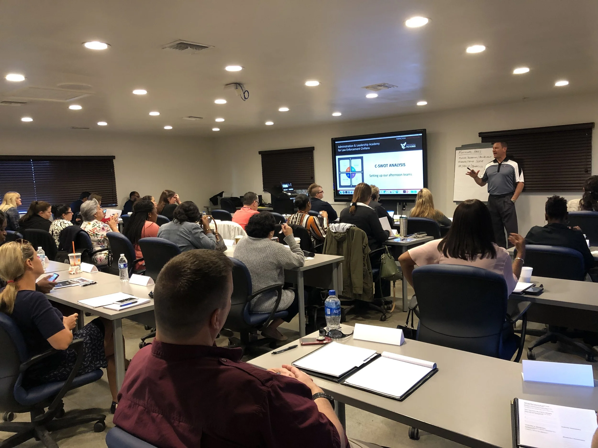 A classroom or conference room with many people seated at tables, attending a presentation. The presenter is standing near a large screen displaying a slide labeled 'C-SWOT Analysis.' Some attendees are taking notes, and there are water bottles, notebooks, and cups on the tables.