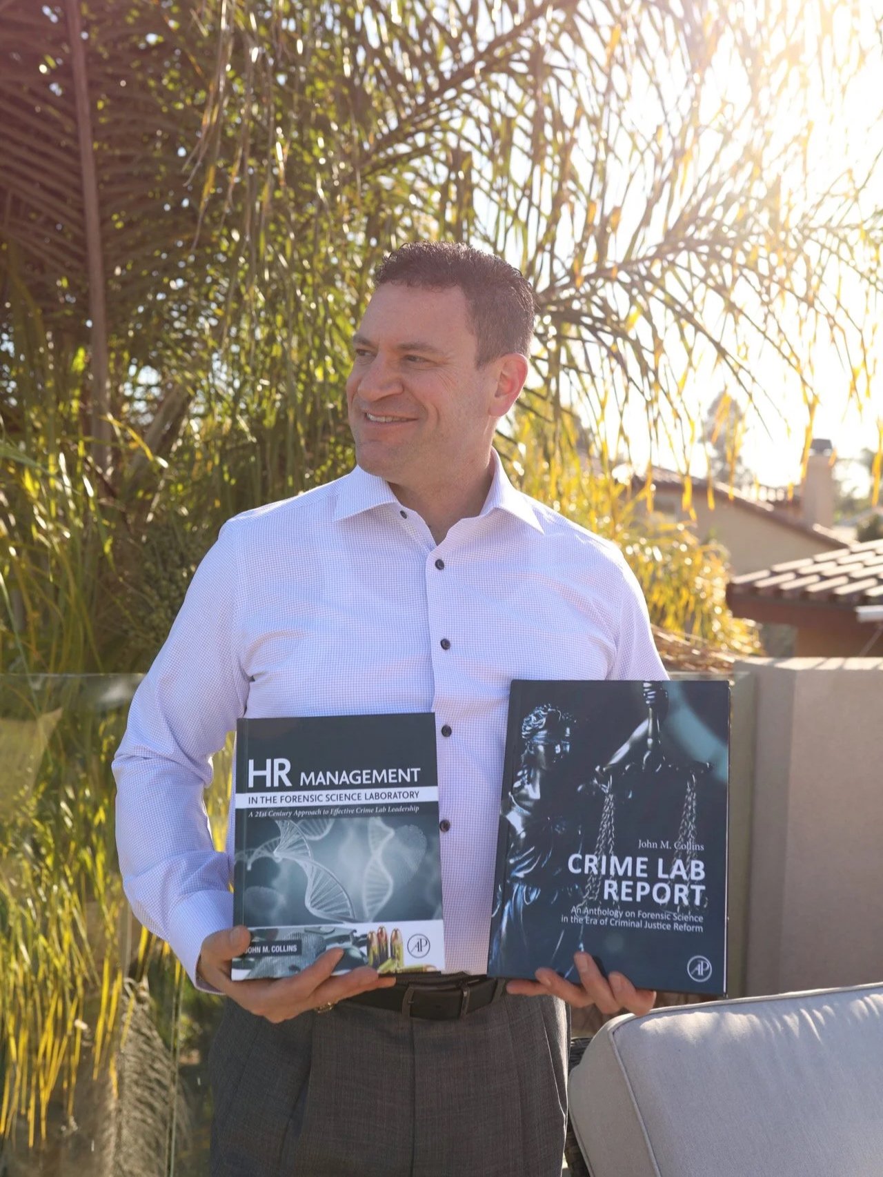 A man in a white dress shirt holding two books about forensic science outdoors on a sunny day.