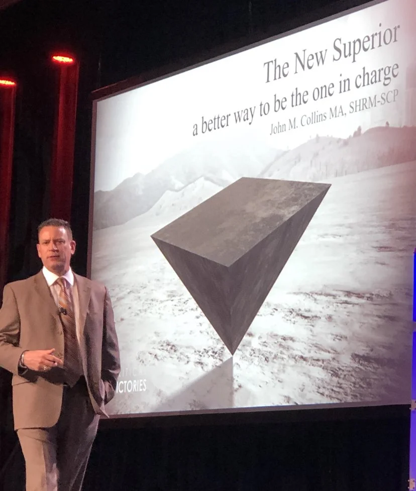 Businessman in a suit stands in front of a large presentation slide on a screen, which displays a large geometric cube and the text "The New Superior: a better way to be the one in charge" by John M. Collins MA, SHRM-SCP. The background of the slide appears to be a barren landscape with mountains.